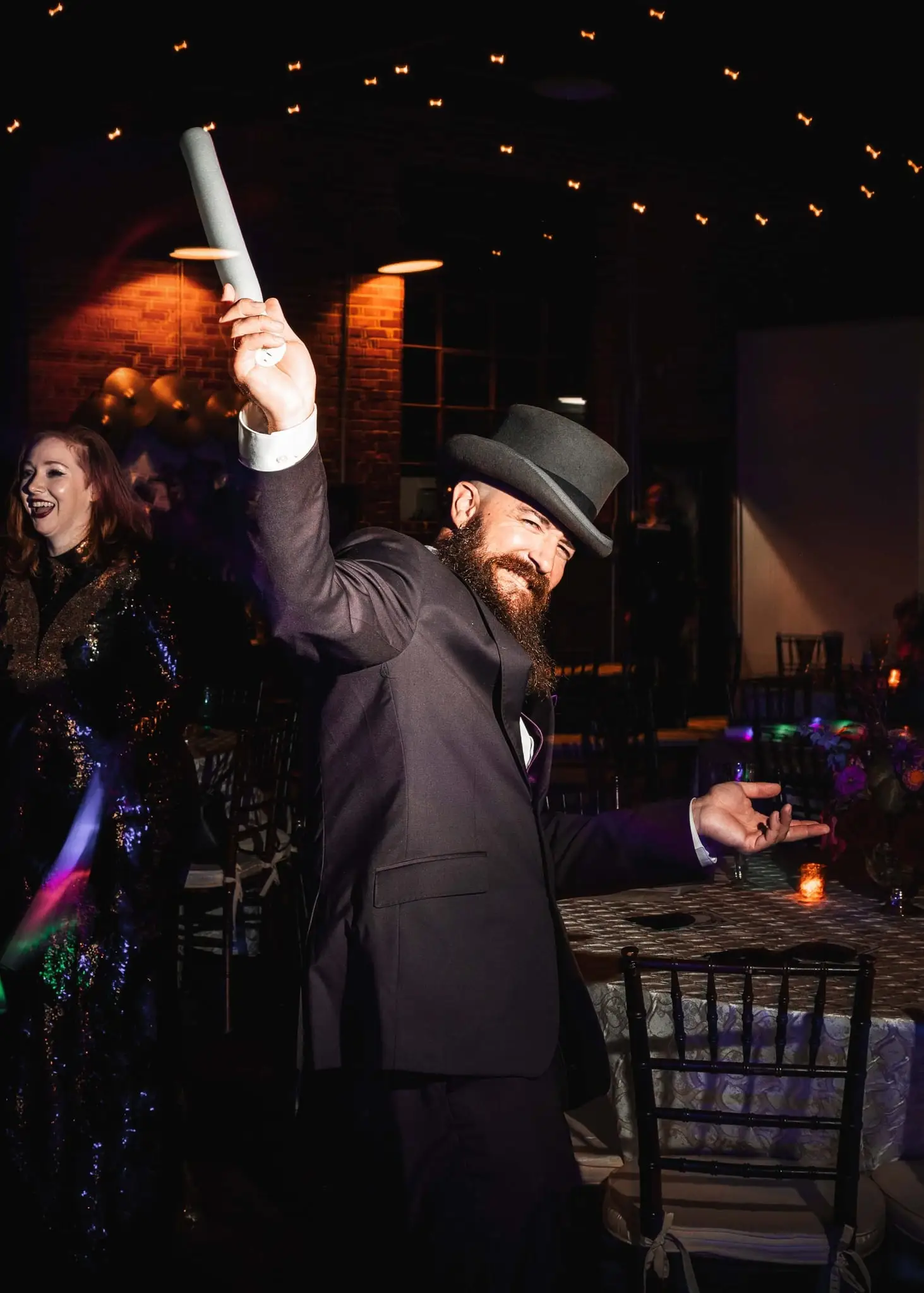 Man in a tuxedo and top hat raising a tube overhead at a reception under string lights