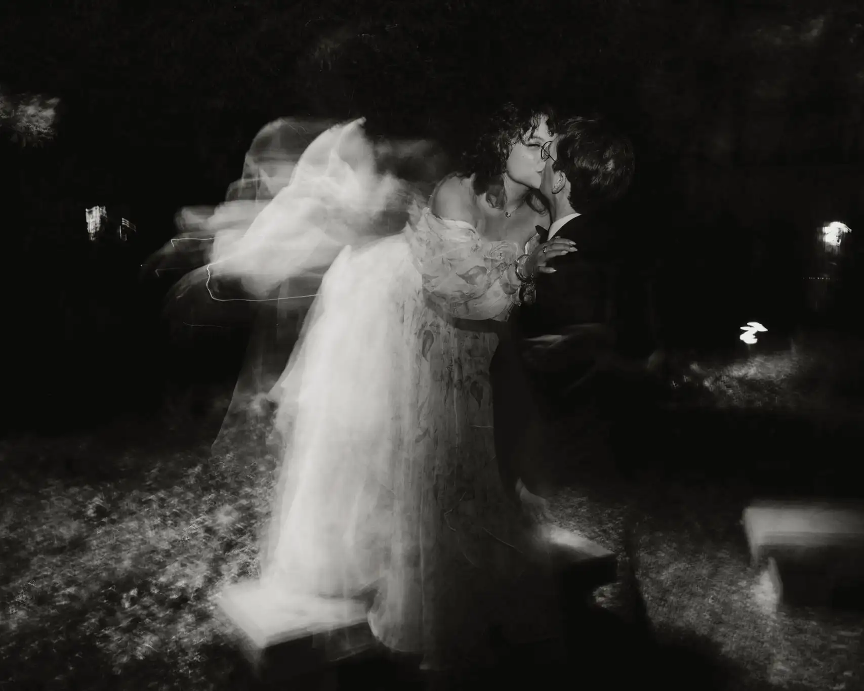 Black and white ghosted long exposure of the bride kissing the groom at night, her gown blurred with motion