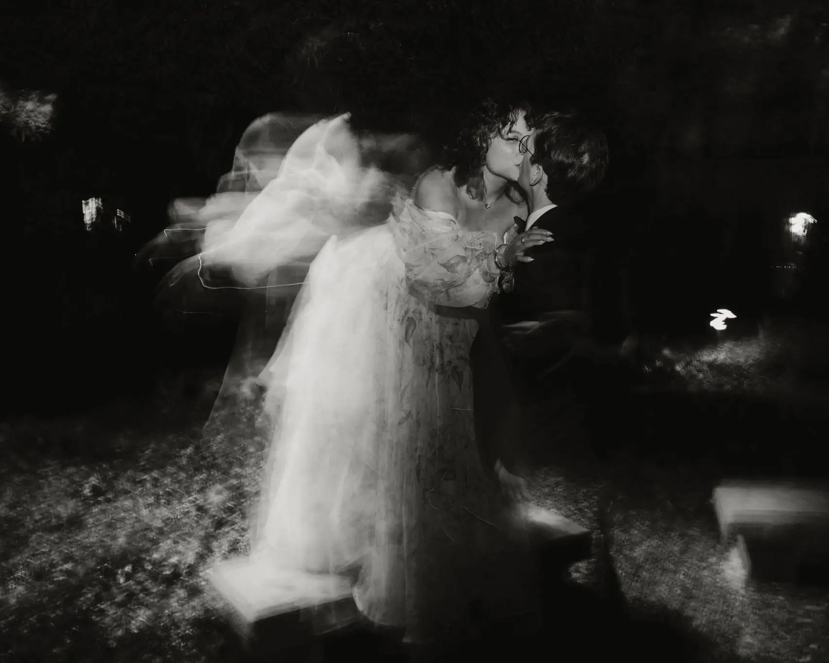 Black and white long-exposure of a couple kissing at night, their motion blurred like a ghost