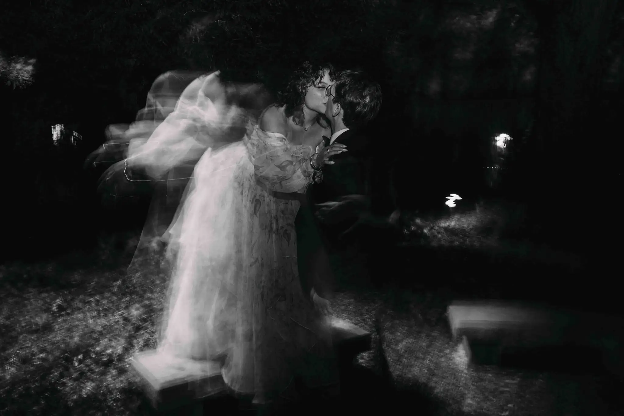 Black and white long-exposure of a couple kissing at night, their motion blurred like a ghost