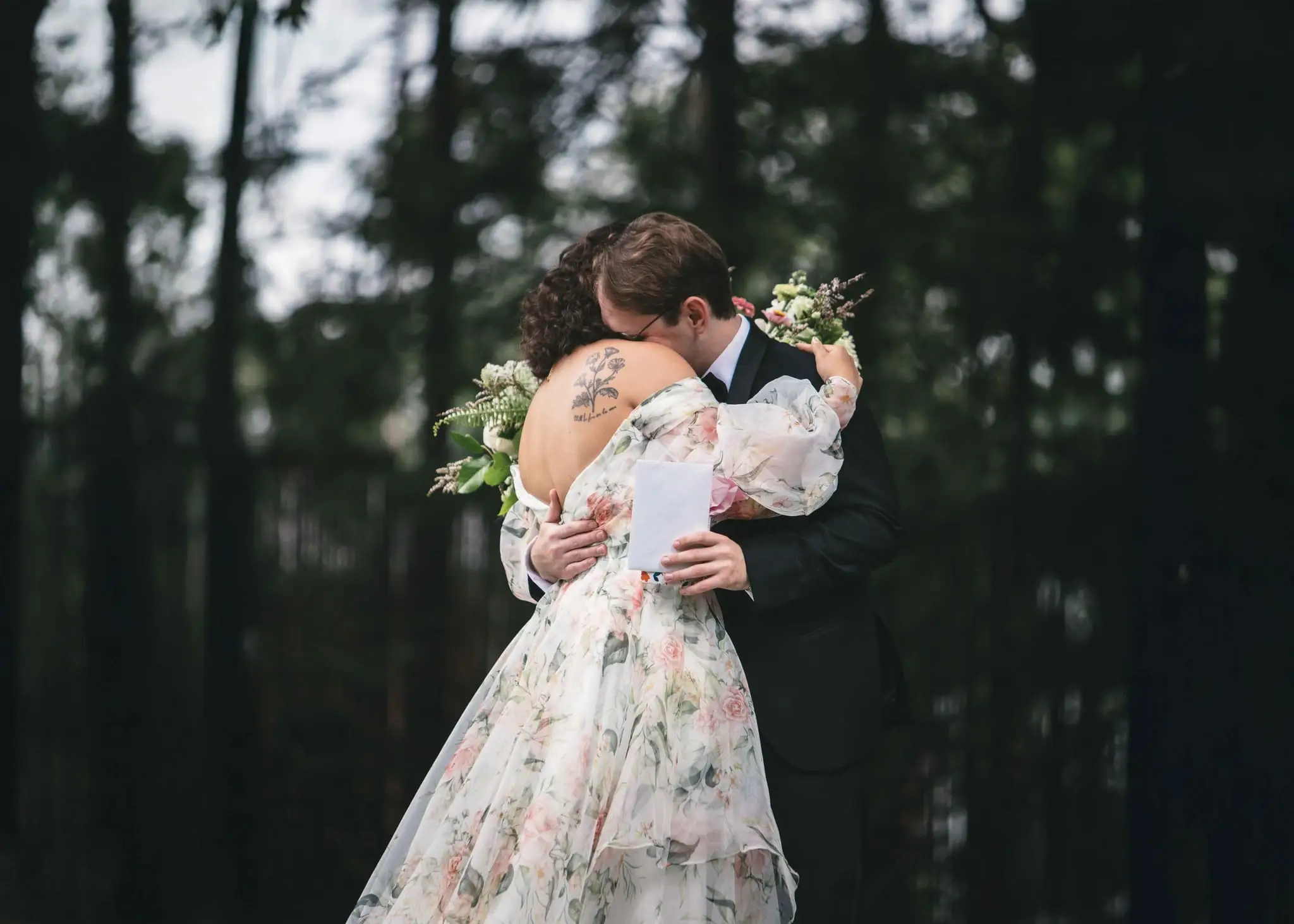 Bride in a floral gown hugging the groom in a pine forest, holding a folded note and bouquet