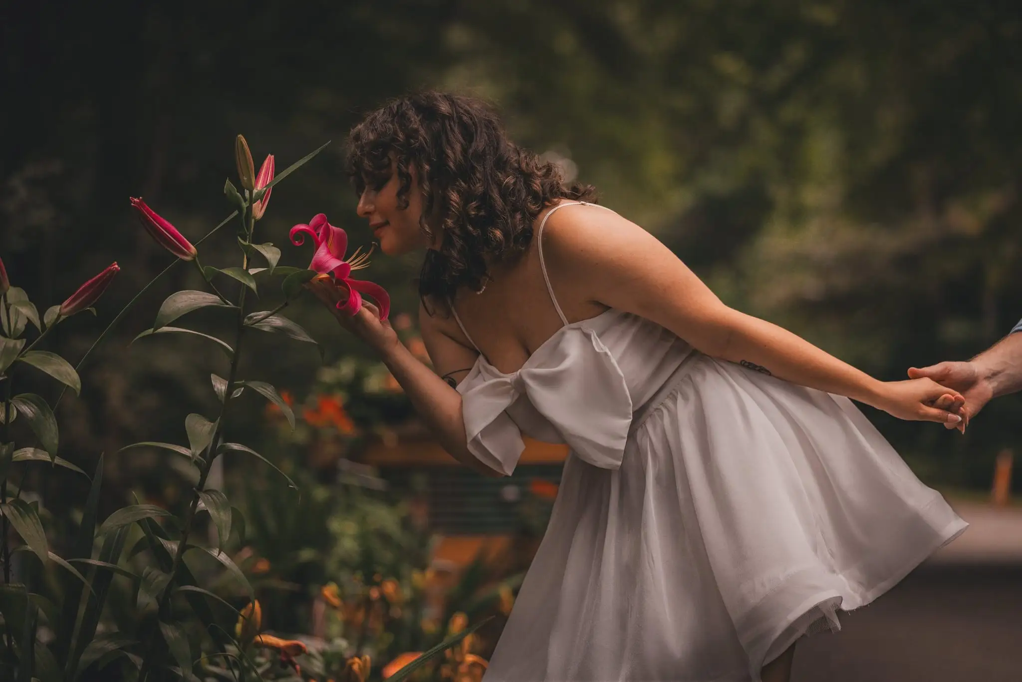 A woman in a short white dress leaning in to smell red flowers while holding her partner’s hand