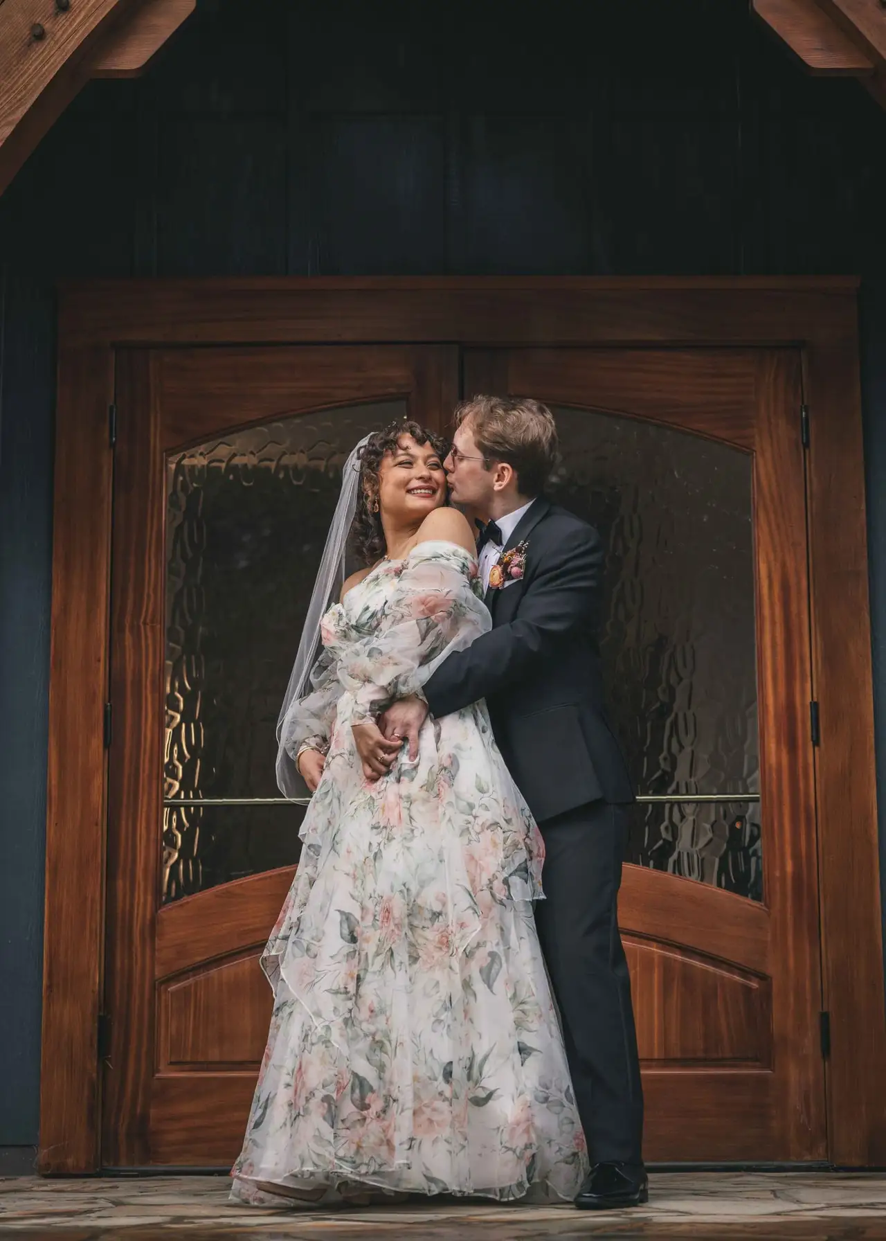 Maya and Jeff wedding at Weaver House, Weaverville NC — the couple kissing in front of a carved wooden double door, the bride in a floral gown