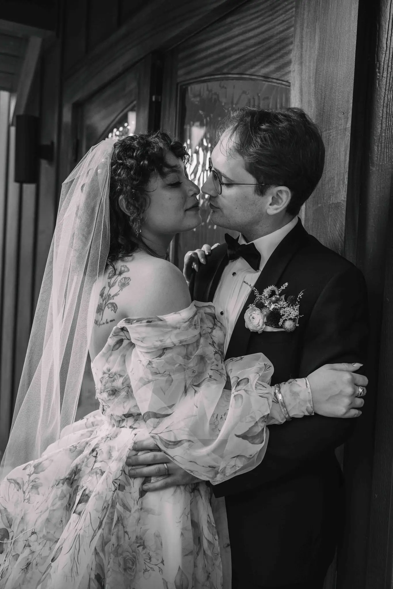 Black and white portrait of the bride and groom almost kissing in a wooden doorway, her veil flowing