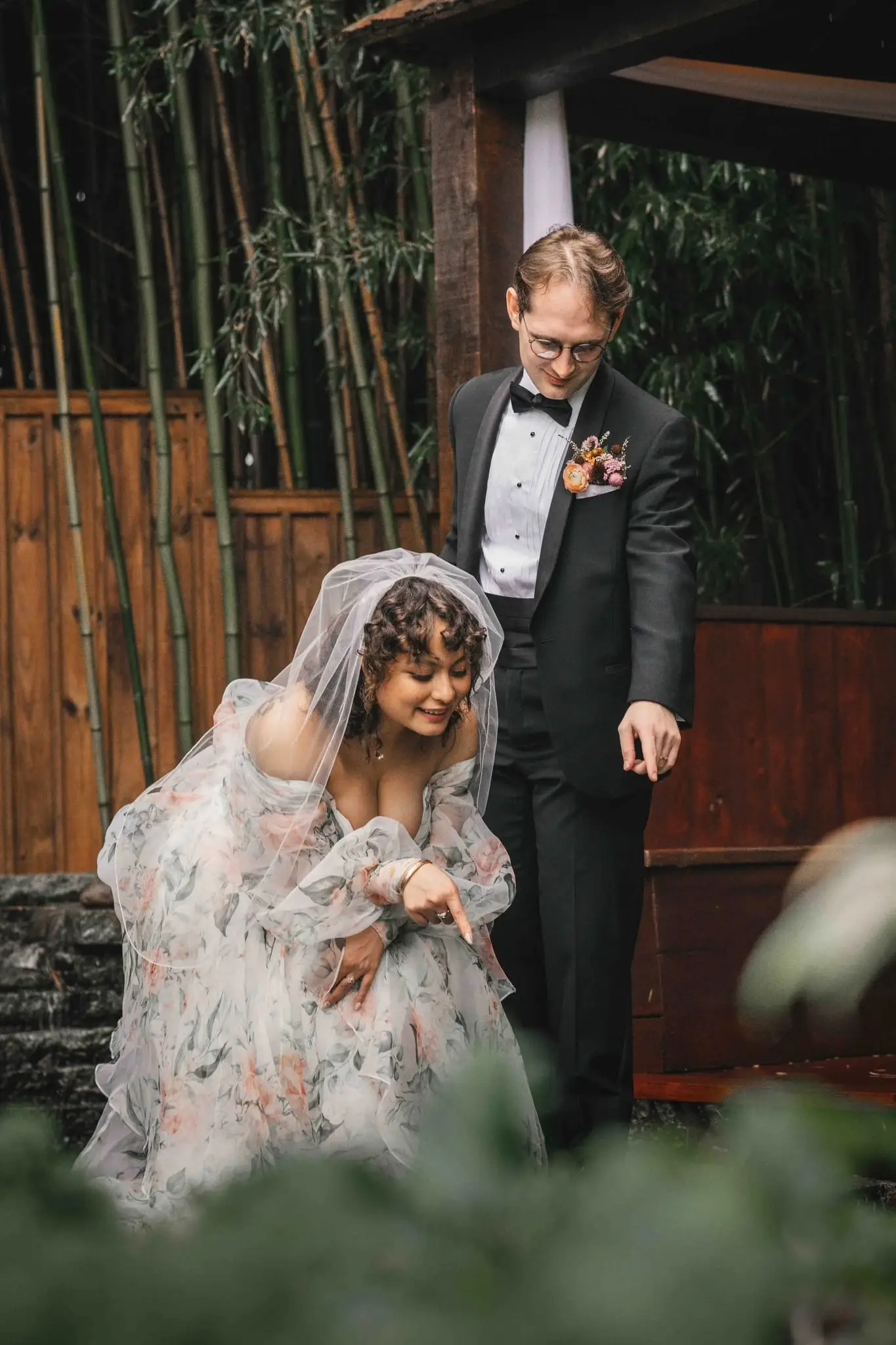 Bride in a floral organza gown and veil pointing at the ground as the groom bends to look, in a bamboo garden