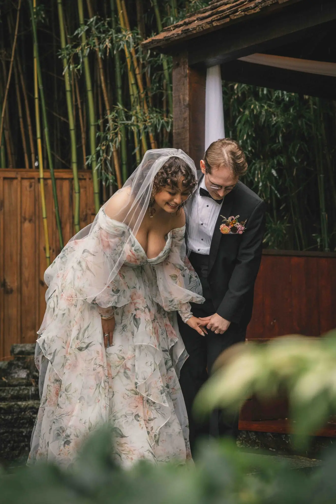 Bride and groom laughing and looking down in a bamboo garden beside a wooden pavilion