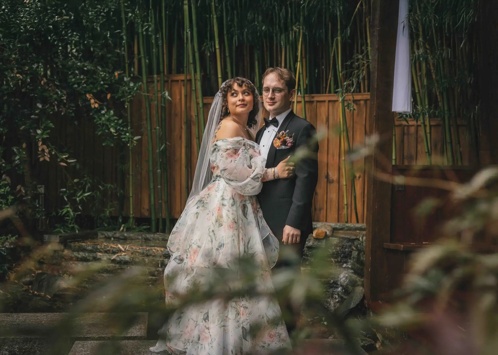 Maya and Jeff wedding at Weaver House, Weaverville NC — the couple standing together in front of a bamboo fence, the bride in a floral gown with a veil
