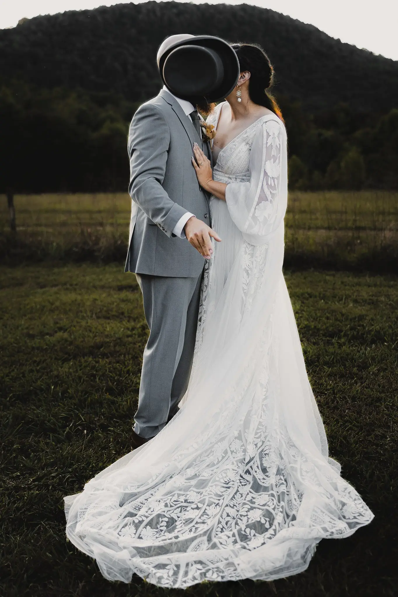 Groom hides a kiss behind his top hat as the bride's lace train spreads across the grass