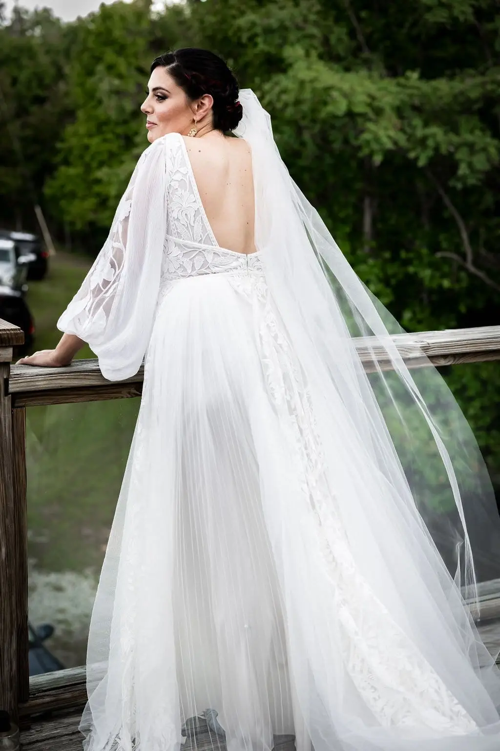 Bride in a flowing veil and lace gown leaning on a wooden deck railing and glancing back