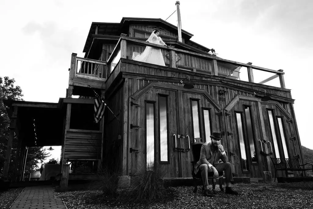 Black and white of the bride on a high wooden deck above the groom seated by the entrance of the rustic house