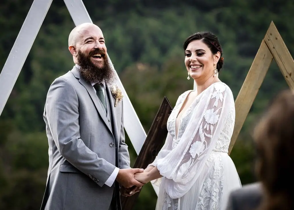 Groom laughing and bride beaming as they hold hands in front of a wooden triangle arch