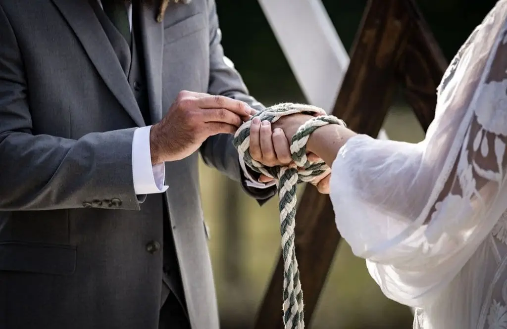 Two people participating in a handfasting ceremony, exchanging vows with hands tied together with a braided rope. One person is wearing a suit, and the other is in a lace dress.