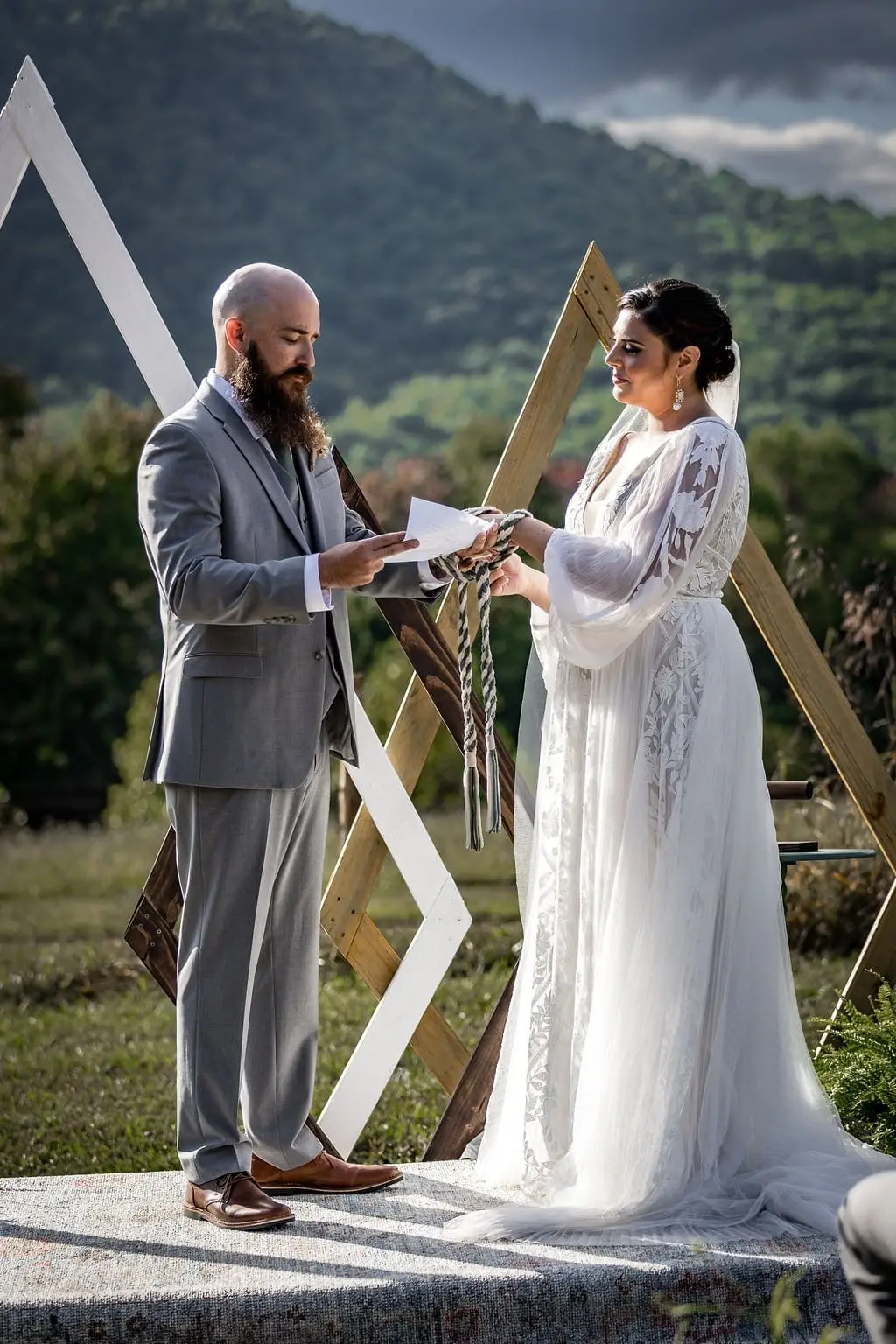 Groom reading from a folded paper to the bride at the altar with a green mountain behind