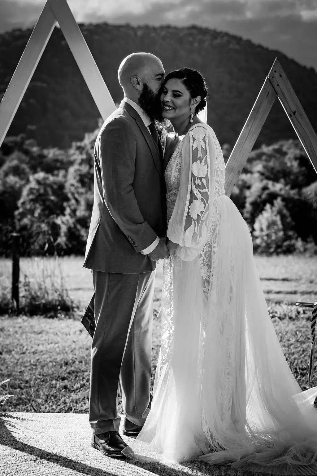 Black and white of the couple embracing cheek to cheek before the arch and mountain ridge