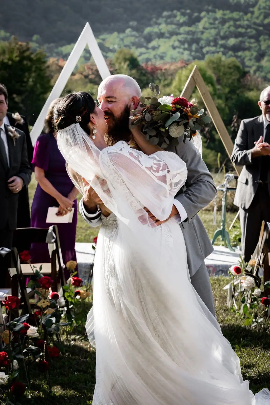Newlyweds kiss in the aisle between red floral arrangements as guests look on