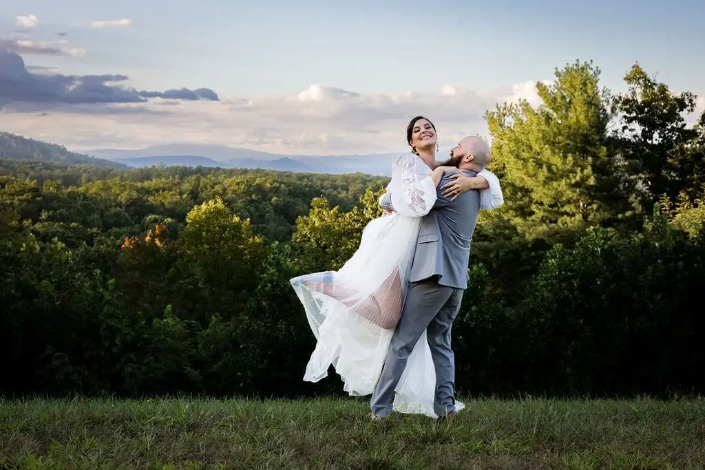 Groom lifting the bride in a joyful hug on a hilltop with sunset-tinted mountains behind