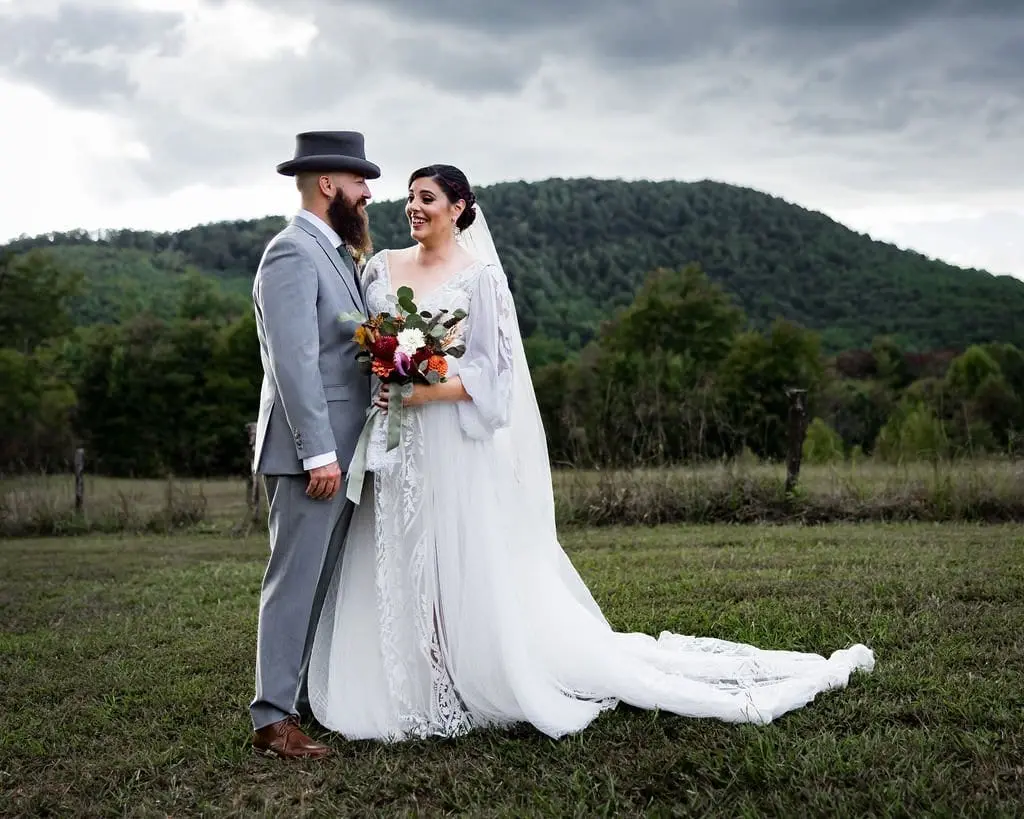 Couple embracing in a mountain-ringed field, the bride holding a fall-toned bouquet under storm clouds