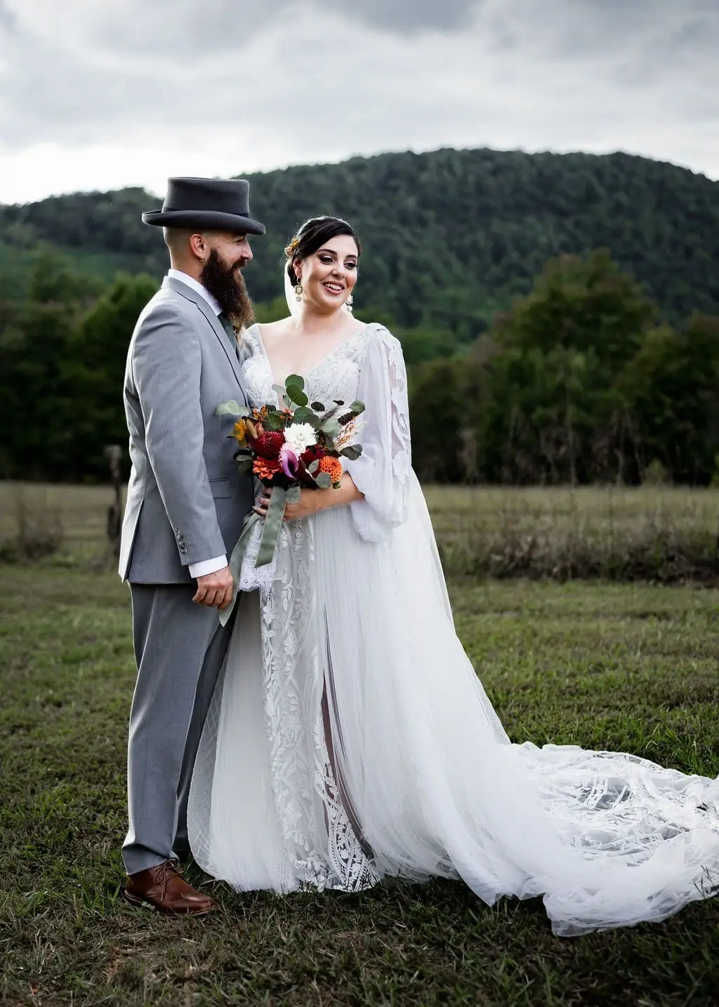 Bride with a colorful bouquet and groom in a top hat in a green pasture under cloudy skies