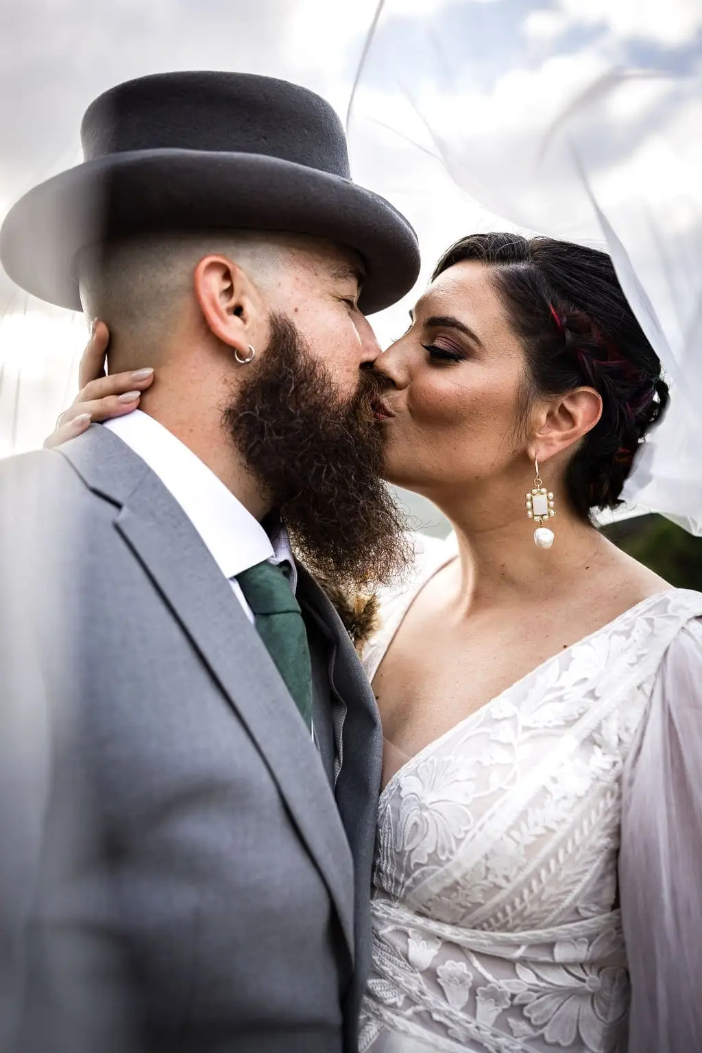 Megan and Brendan kissing outdoors, he in a gray suit and black top hat, she in a white lace gown