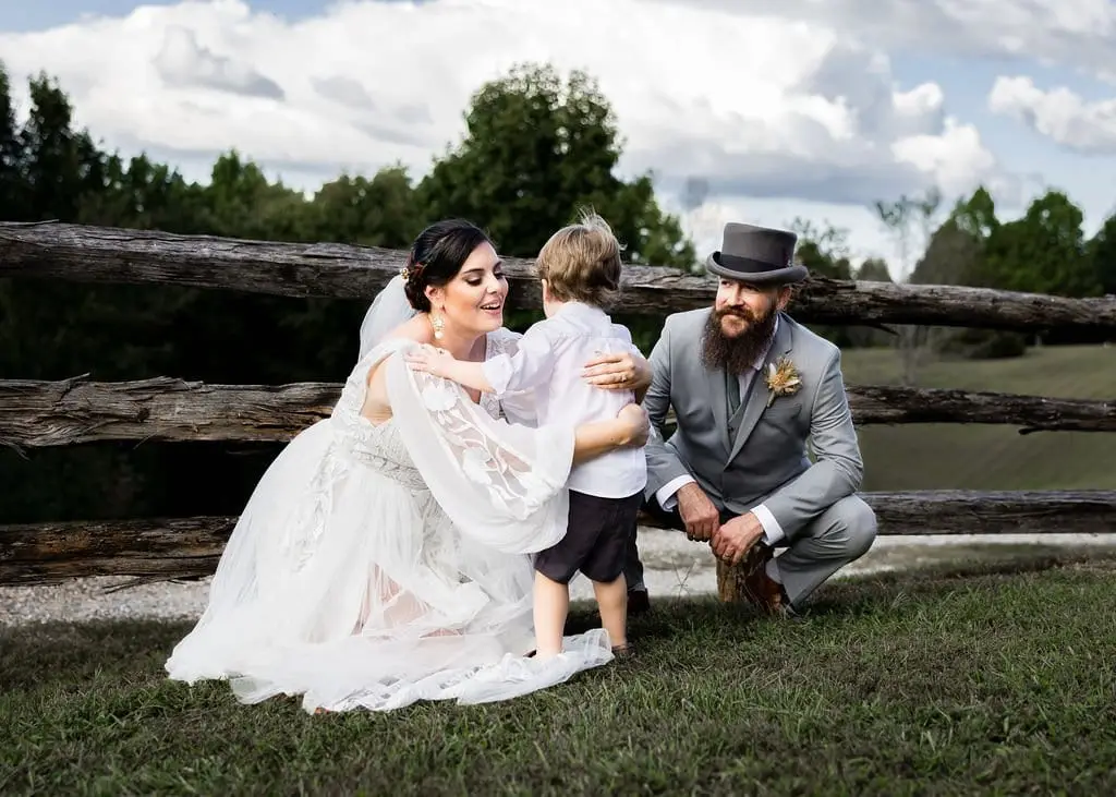 Bride and groom crouching to hug a small boy beside a split-rail fence