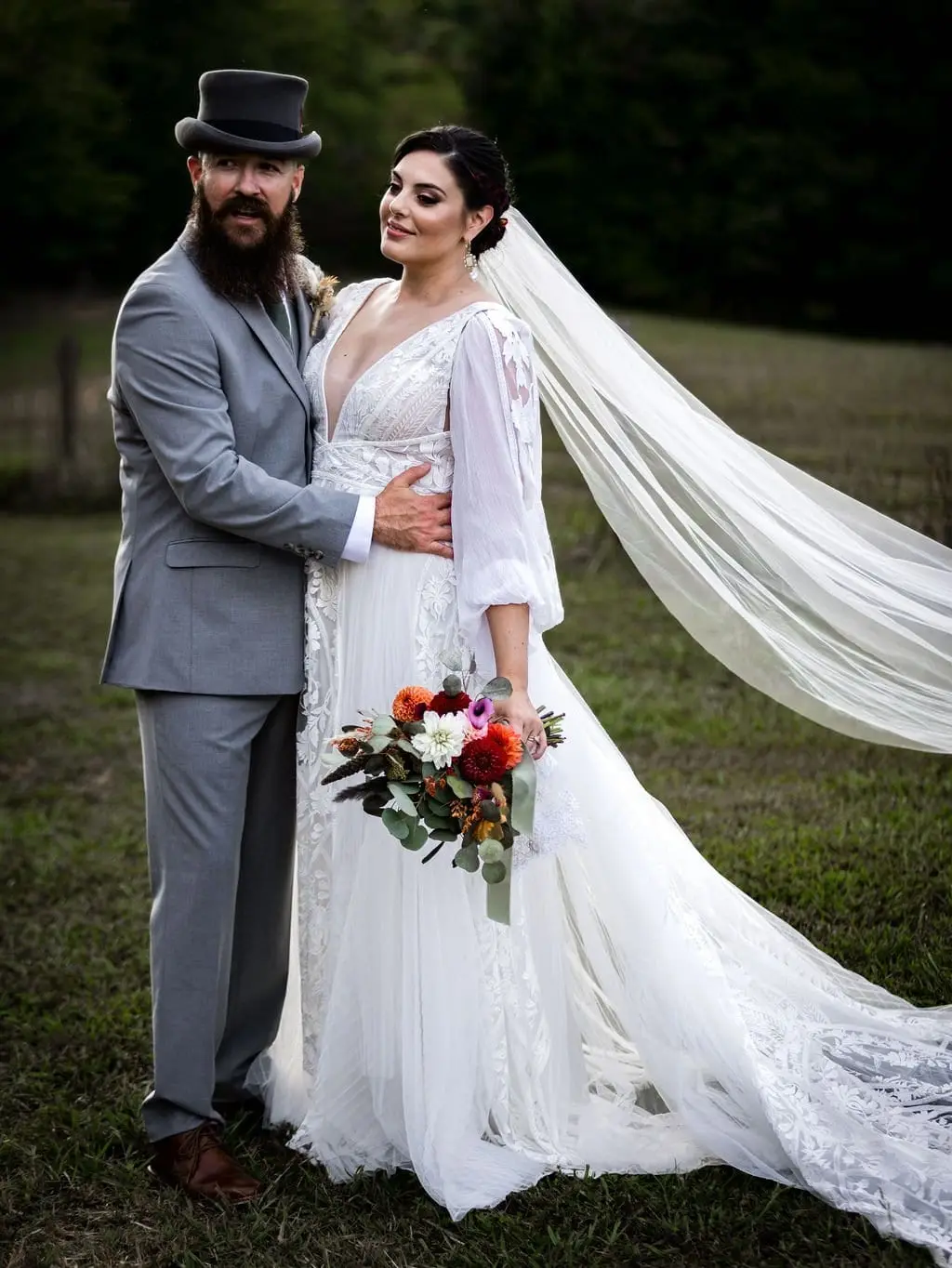 Groom in a top hat holding the bride as her long veil blows, with an autumn-toned bouquet