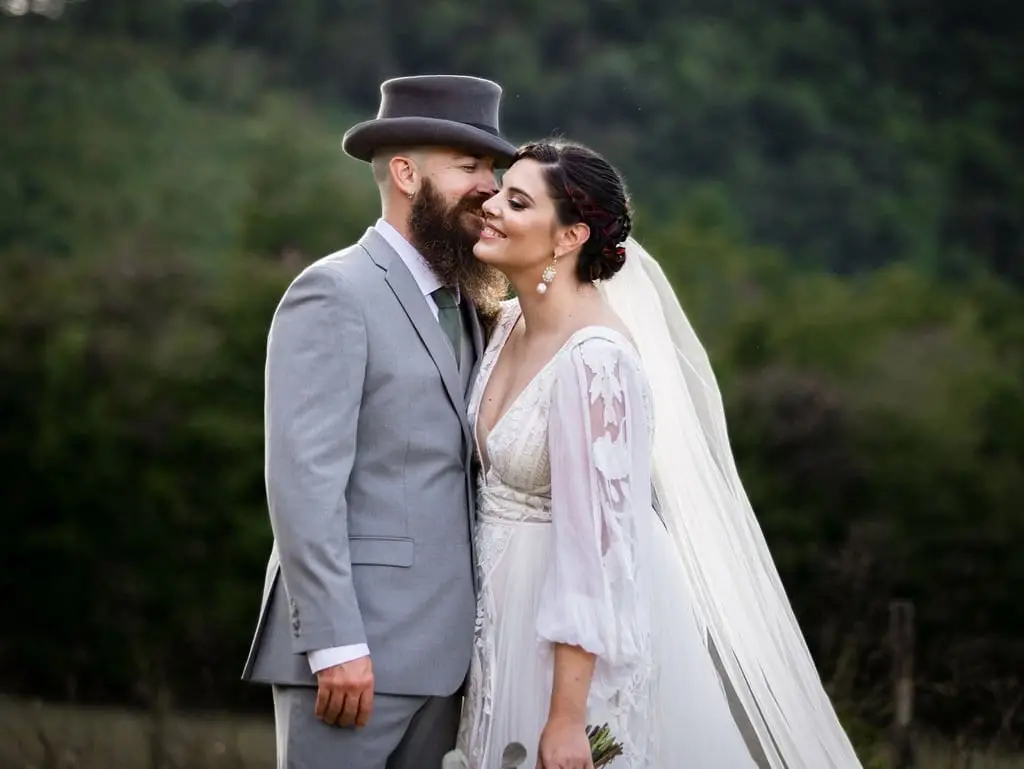Groom in a top hat kissing the bride's cheek as she smiles with her eyes closed