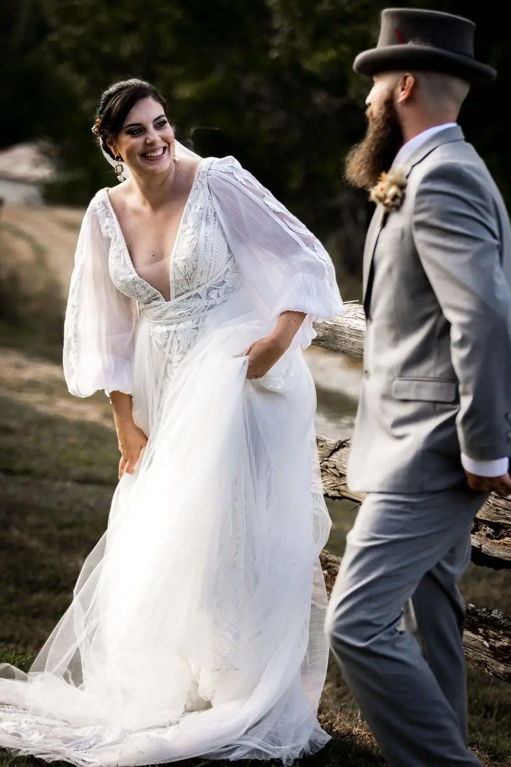 Bride laughing and holding her skirt as the groom in a top hat walks past in golden light