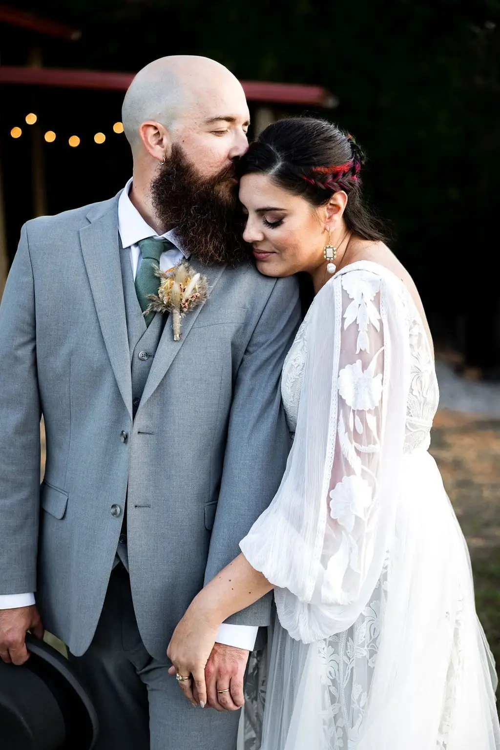 Bride resting her head on the groom's shoulder with string lights glowing behind them