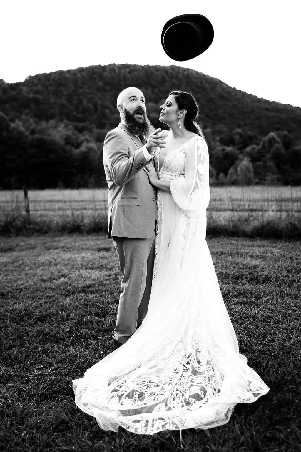 Black and white of the groom tossing his top hat in the air beside the bride and her long lace train