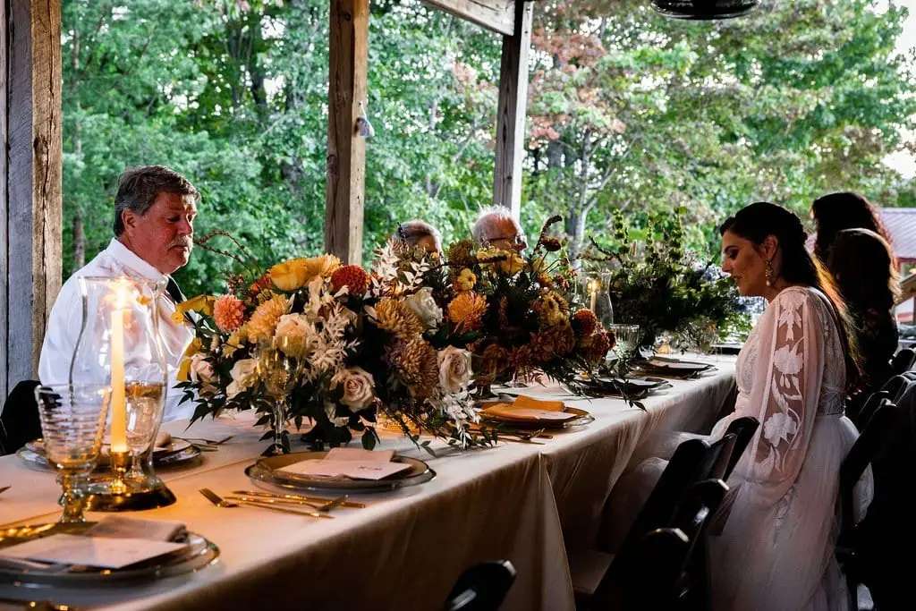 Bride and guests seated at a long table with orange dahlia centerpieces on a covered porch