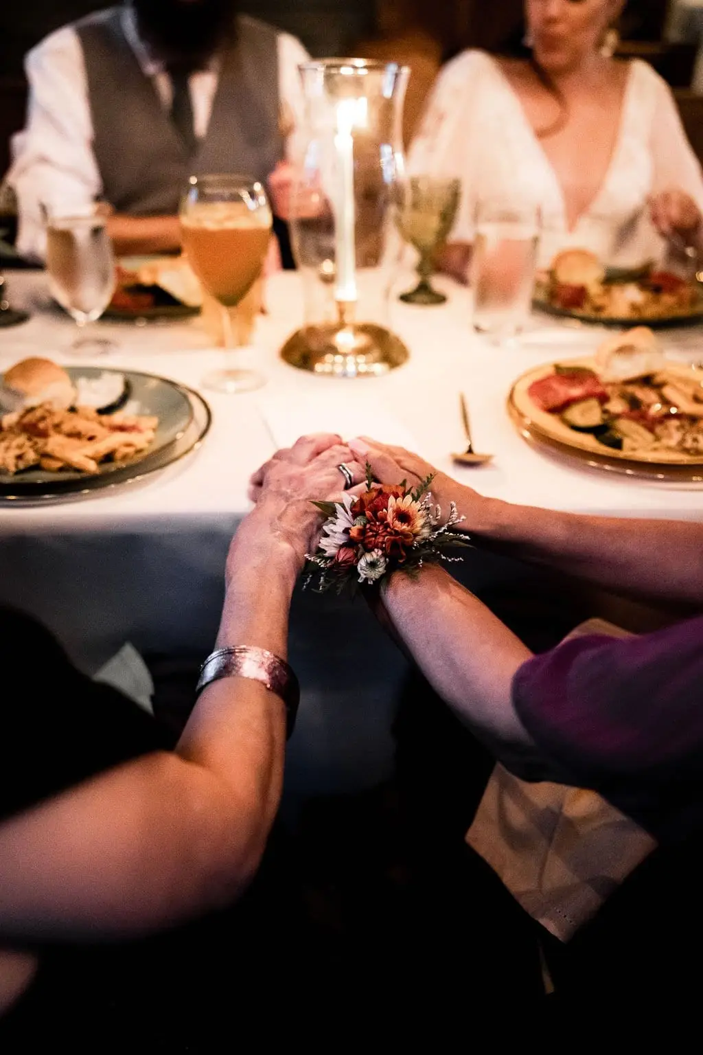 Two guests holding hands, one wearing a flower wrist corsage, at a candlelit dinner table