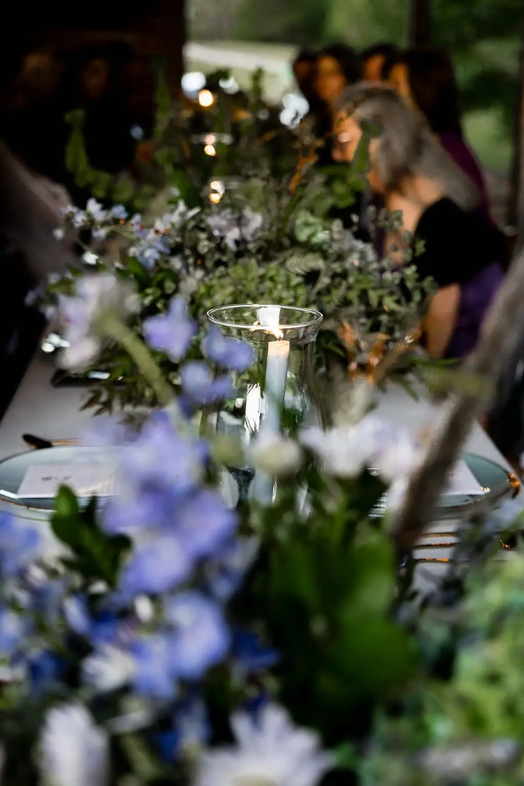 Blue delphinium and greenery centerpieces lining the candlelit reception table with guests beyond