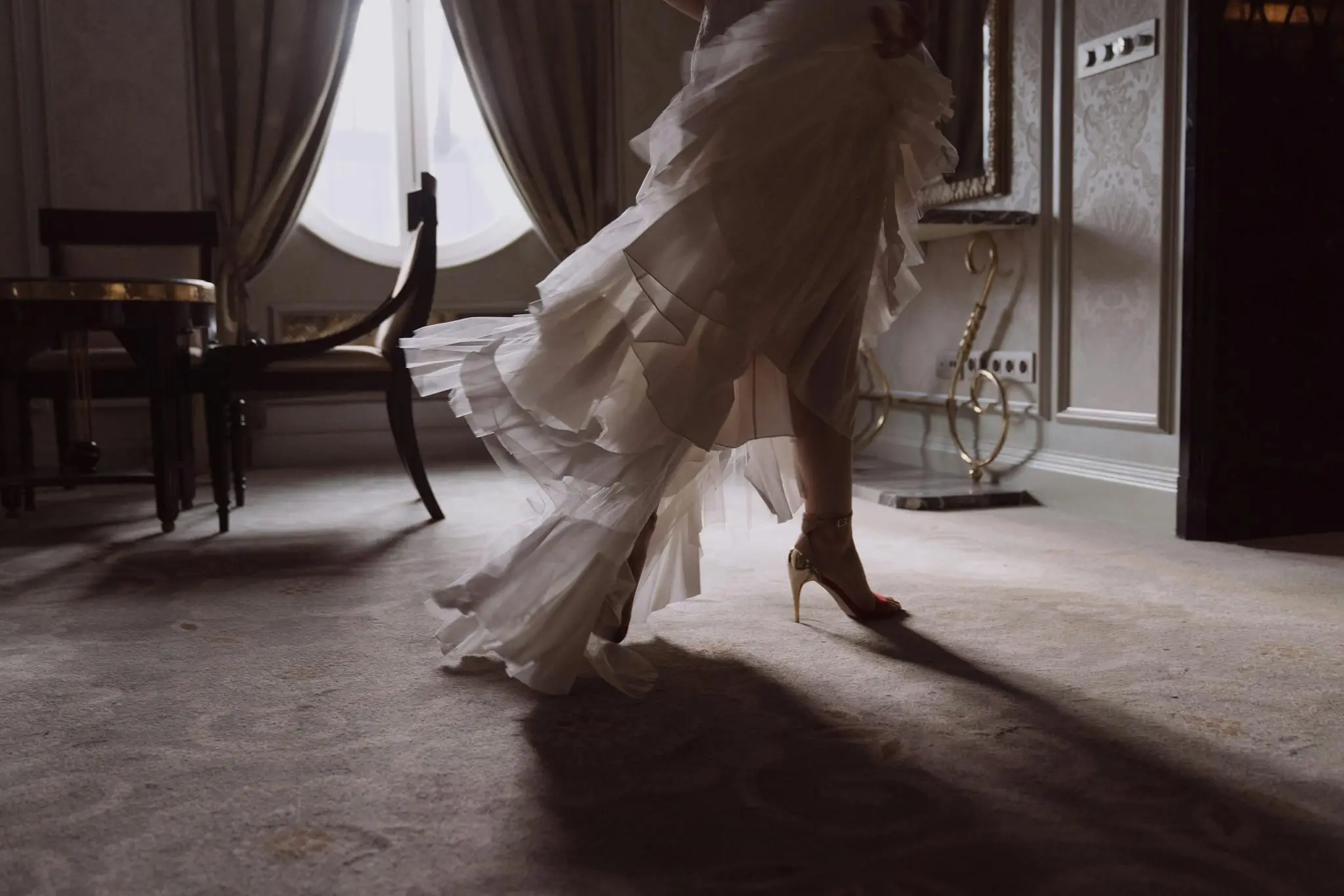 Bride in a tiered ruffle gown and heels crossing a hotel suite, her shadow on the carpet