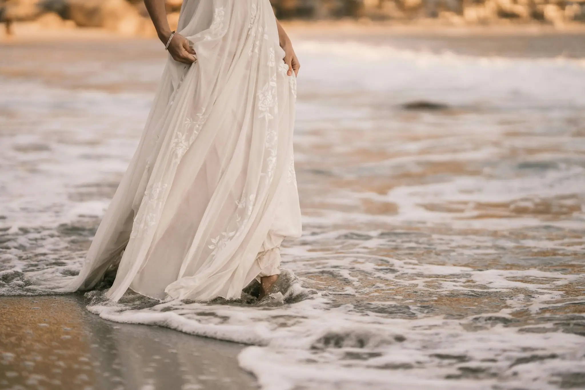 Bride in a lace gown wading barefoot through ocean surf at sunset