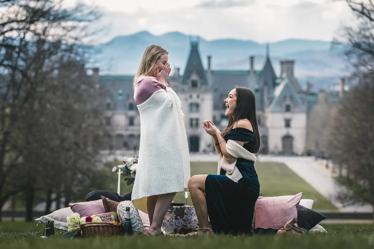 A woman on one knee proposing on the lawn in front of Biltmore House, her partner covering her face in surprise