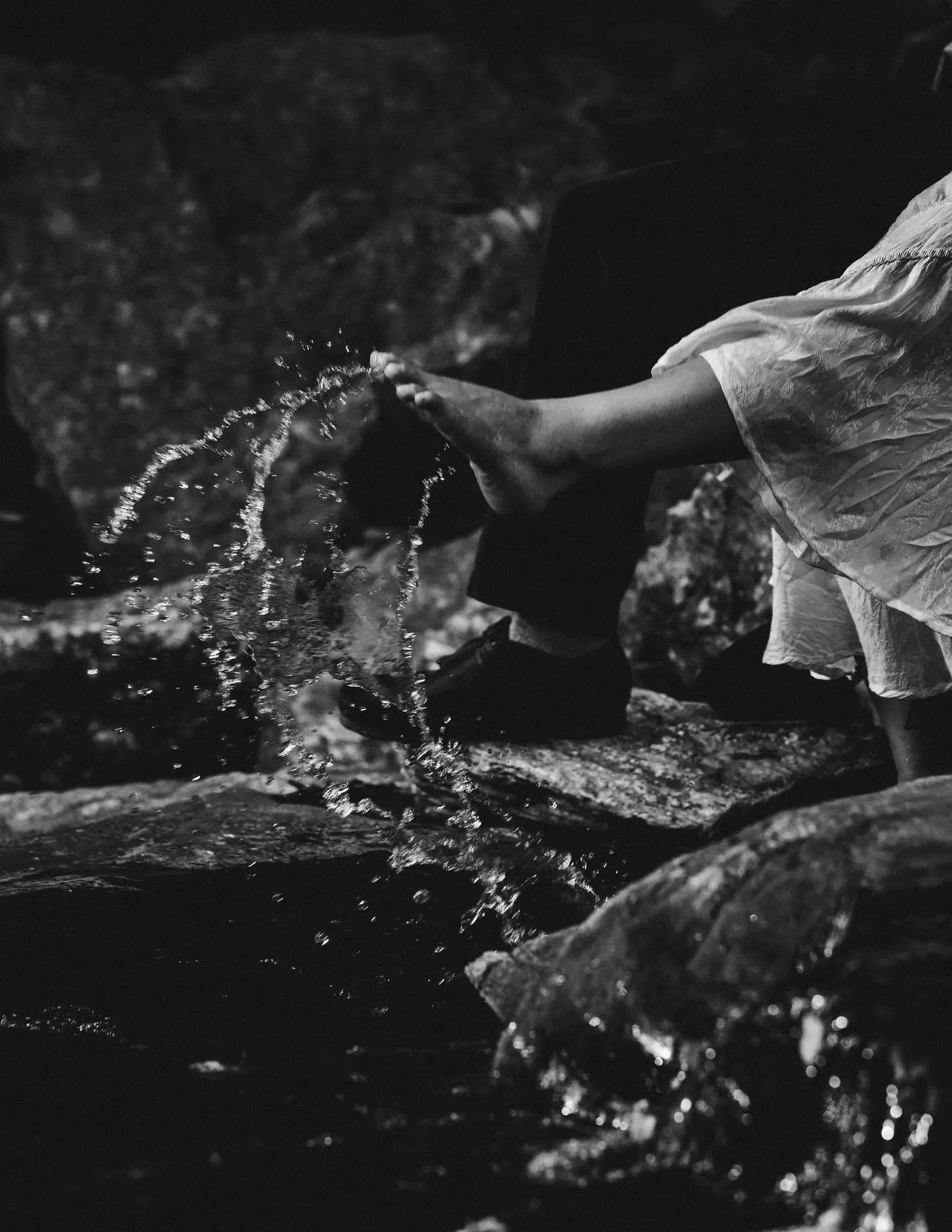Black and white of a bare foot splashing in a creek over dark rocks, a dress hem above