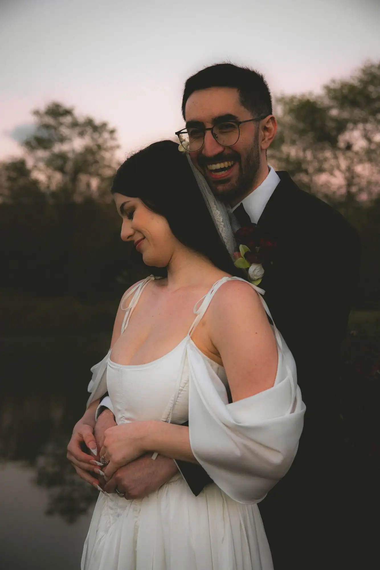 Groom holding the bride from behind at dusk, both laughing
