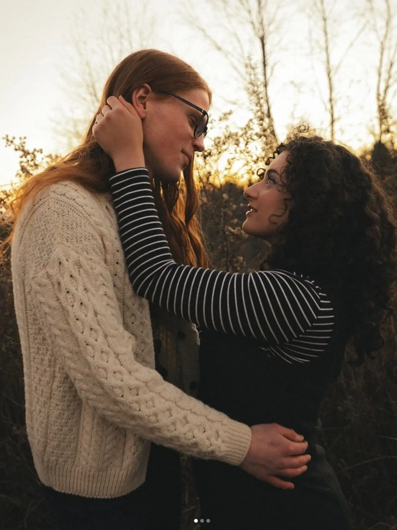 Couple embracing in golden sunset light among tall grass and trees, one hand on the other's face