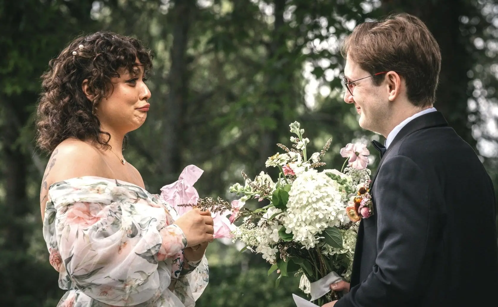 A man in a black tuxedo holding a bouquet of flowers and a woman in an off-shoulder floral dress exchanging a gift outdoors surrounded by green trees.