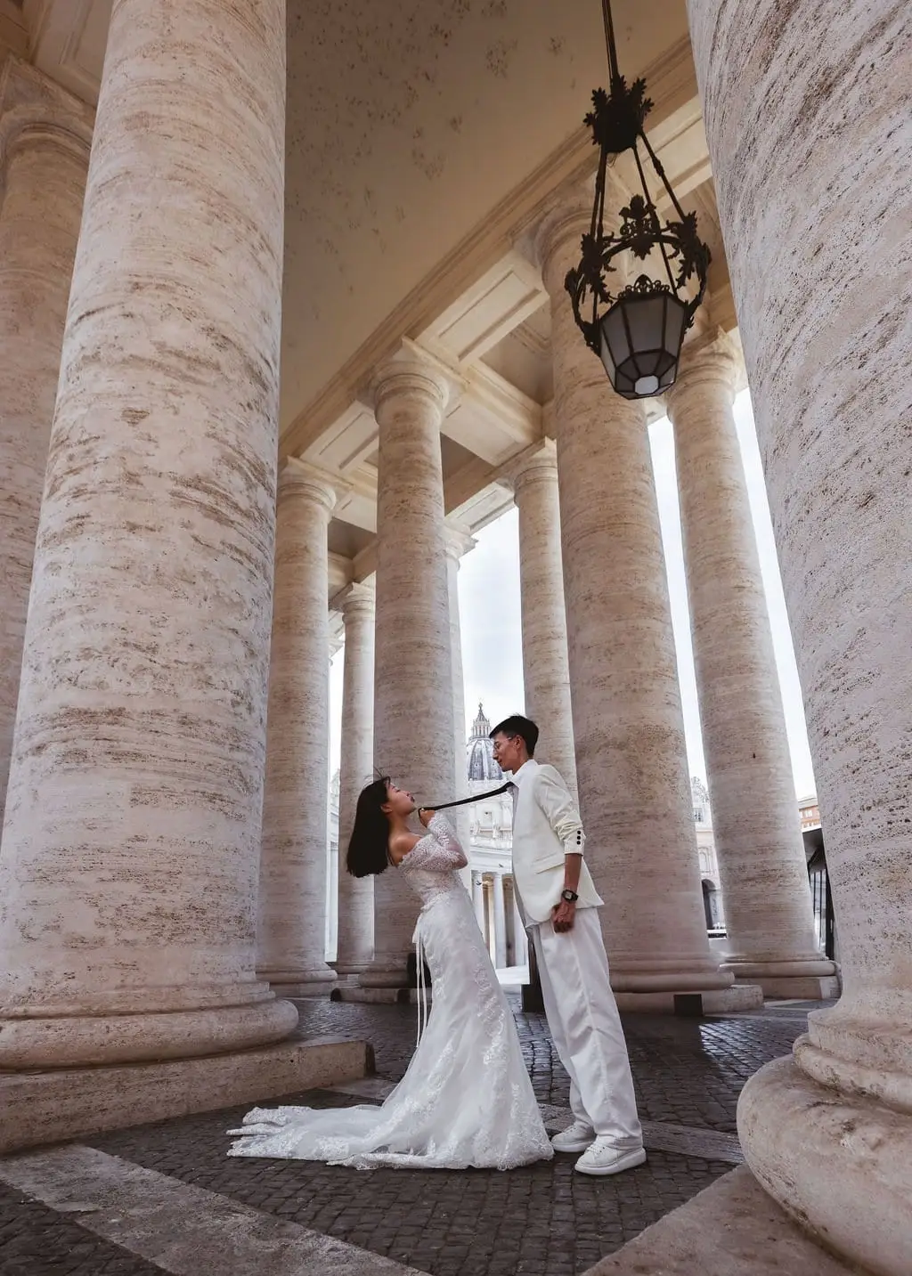 Couple between giant stone colonnade columns, the bride tugging the groom's tie as both laugh