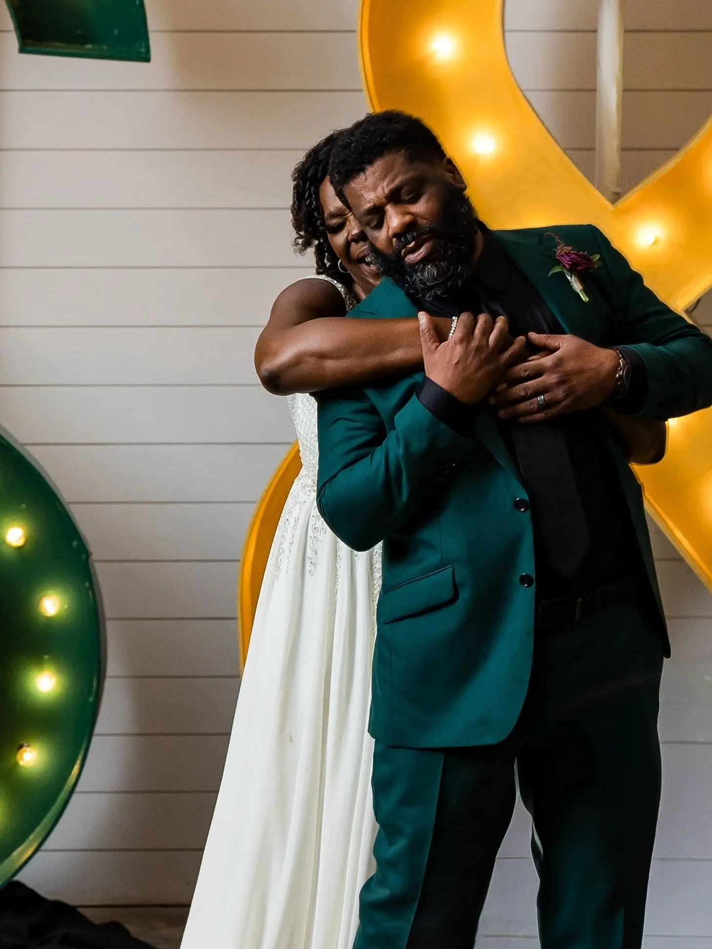 Groom in a teal suit embraced from behind by the bride in front of glowing marquee letters