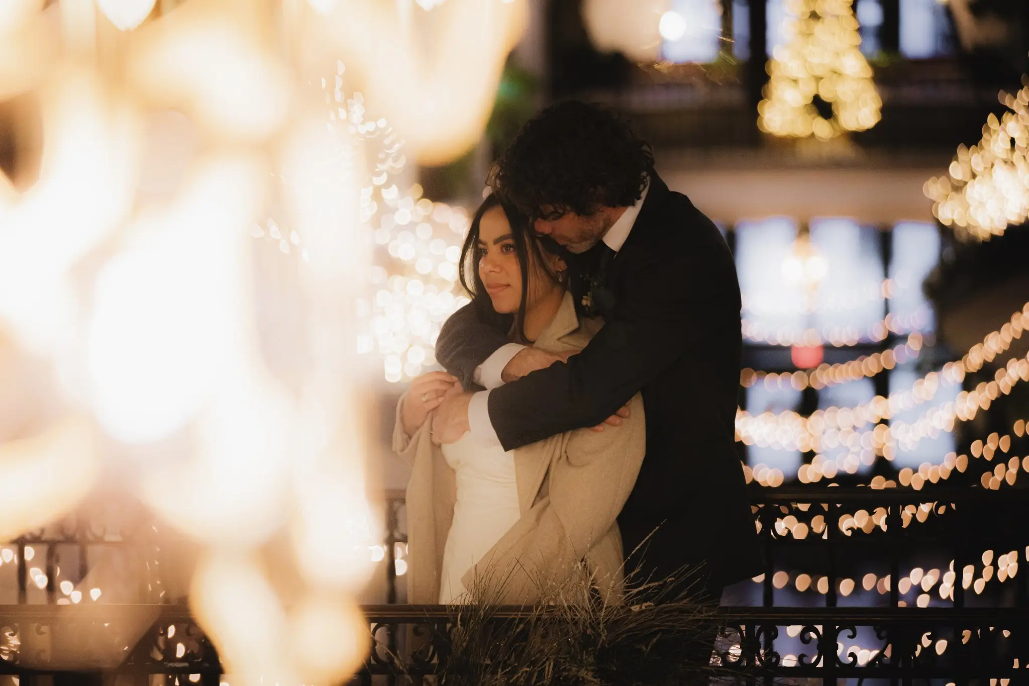 Groom hugging the bride from behind on a balcony glowing with holiday lights