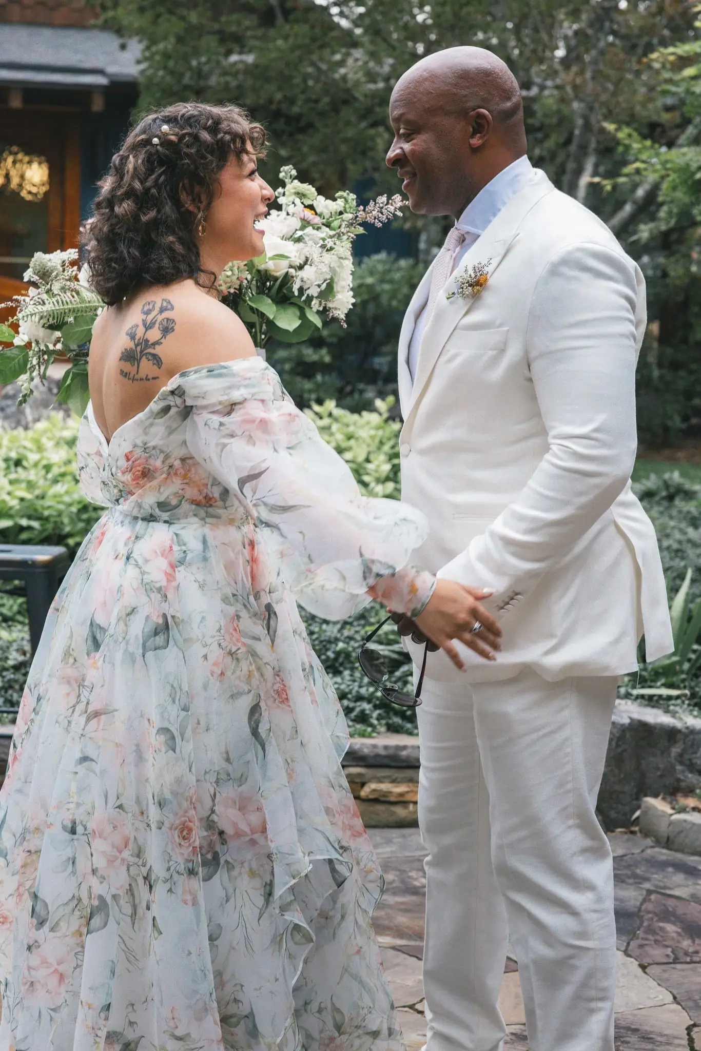Bride in a floral gown laughing with a man in a white suit in a garden