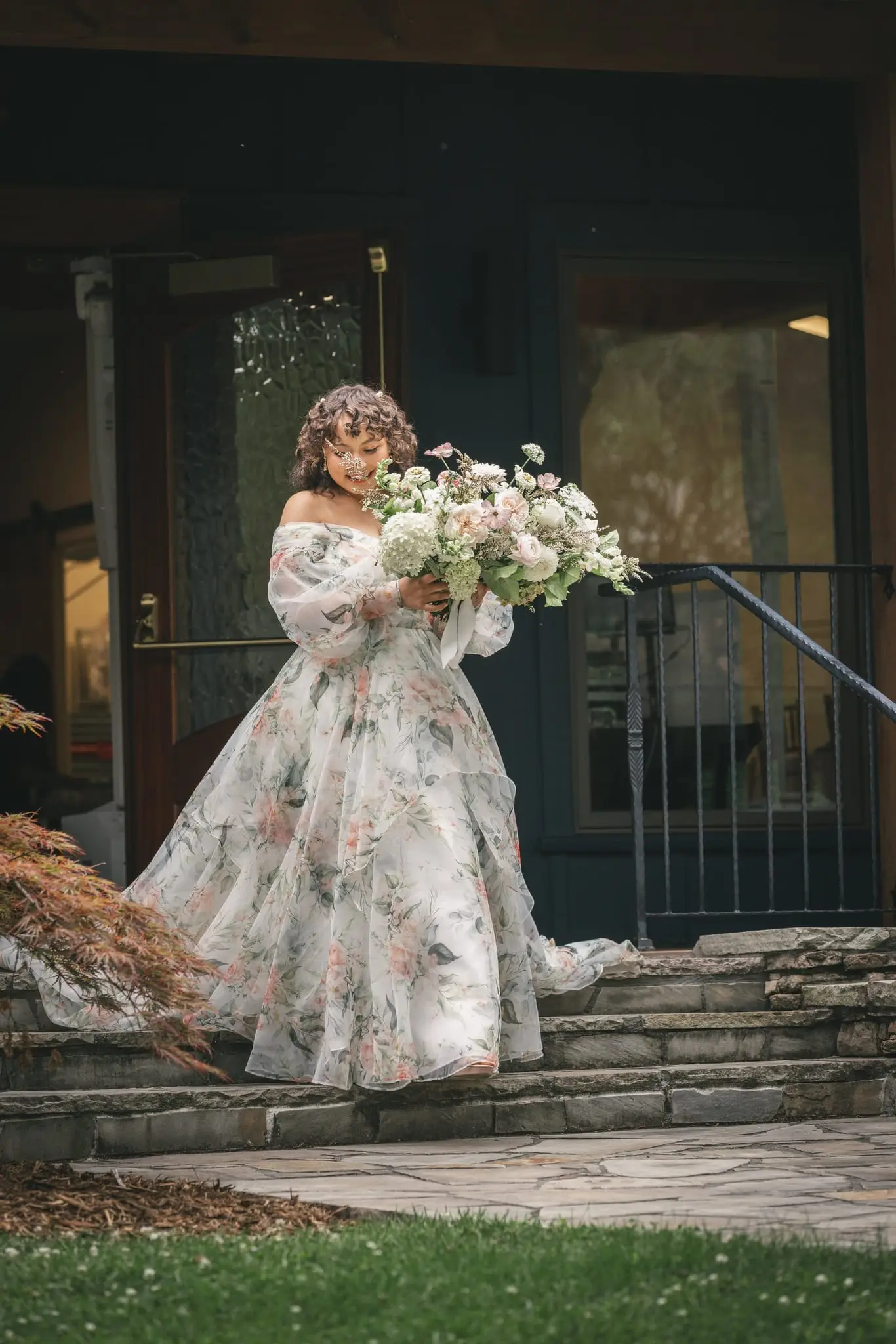 Bride in a floral organza gown holding a bouquet on the stone steps at Weaver House